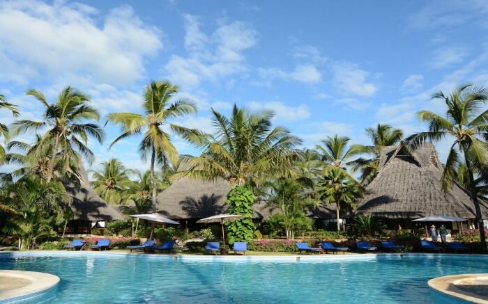 an open pool surrounded by palm trees in a Zanzibari thatched roof resort